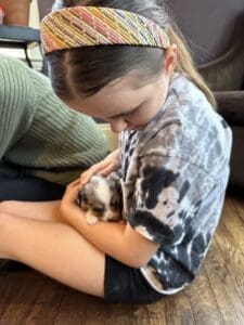 A young girl wearing a tie-dye shirt and a colorful headband sits on a wooden floor, gently cradling a small, fluffy puppy in her arms.